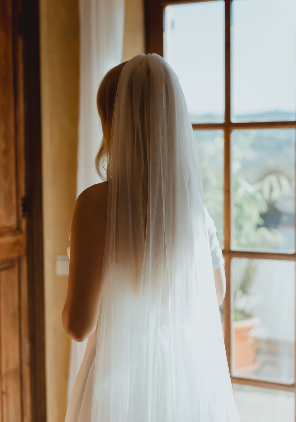 Long white wedding veil flowing against wooden windows in warm indoor lighting.