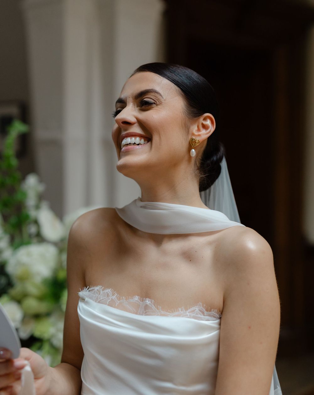 Smiling bride in strapless white wedding gown with veil and gold pearl earrings, laughing joyfully at outdoor ceremony.