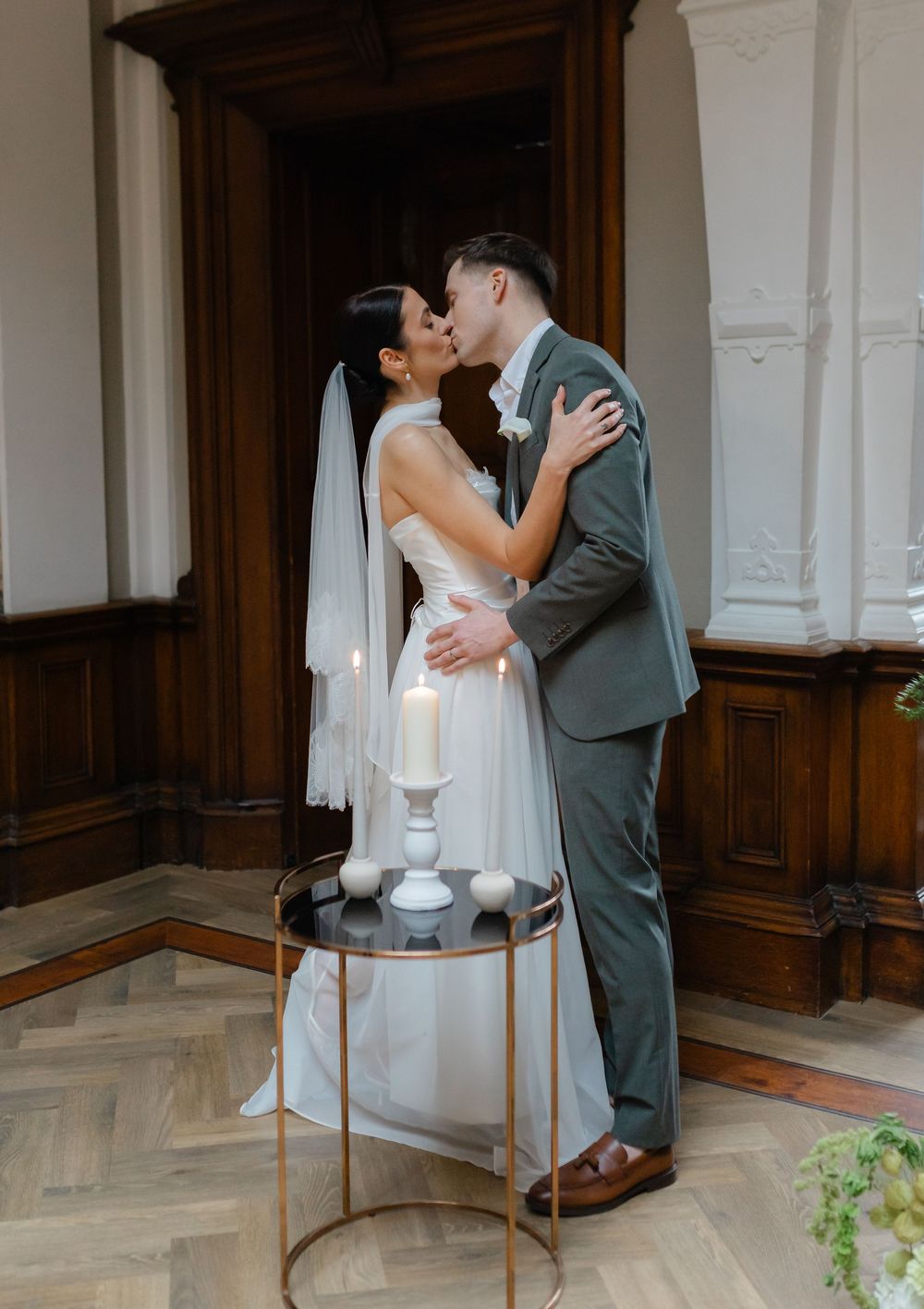 Bride and groom share a first kiss at altar with lit candles on gold stand in elegant wood-paneled venue.