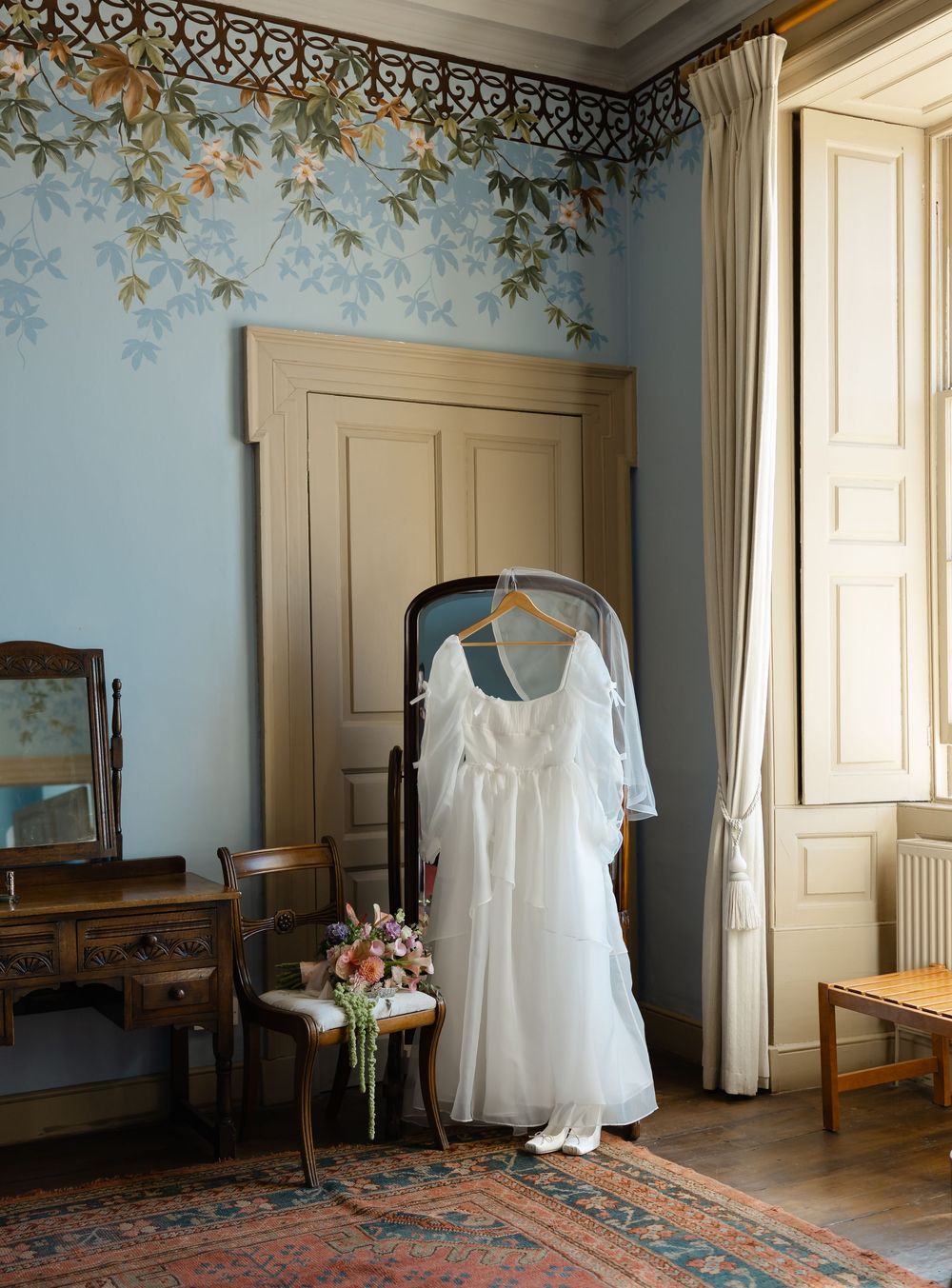 White wedding dress hanging on a chair in an elegant blue room with floral wall murals and antique furniture.