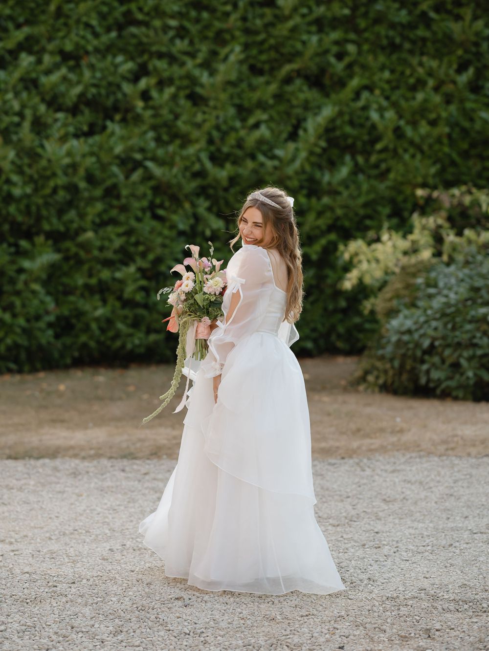 Bride in a white layered tulle gown holding a pink floral bouquet, standing on gravel path with lush green hedges.