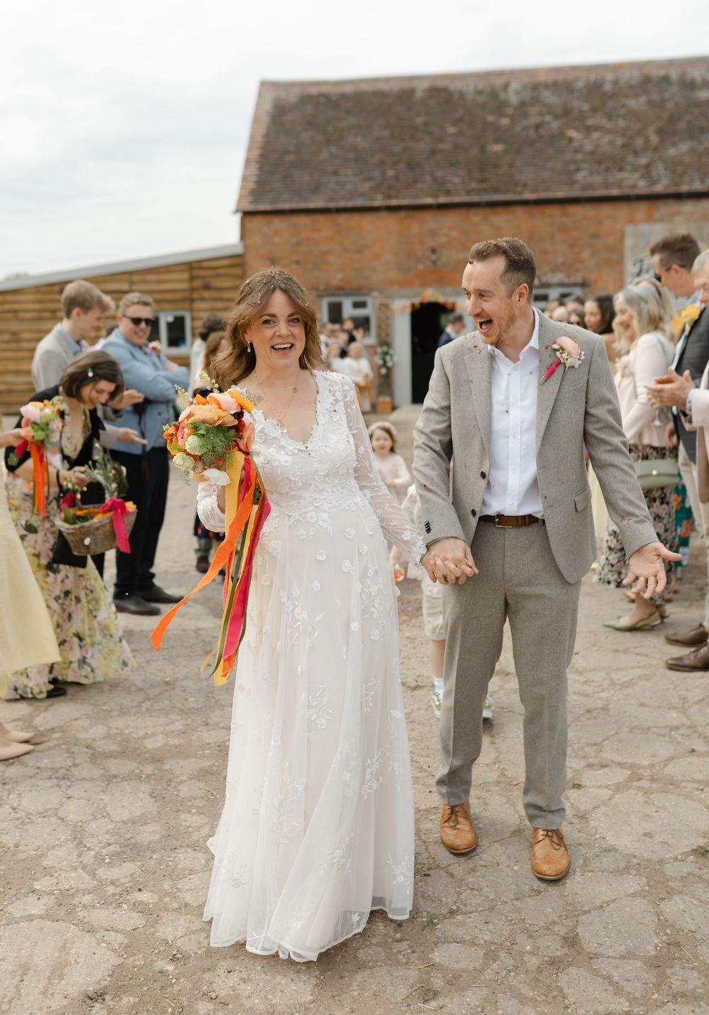 Joyful bride in lace gown and groom in gray suit walk through confetti at rustic barn wedding venue.