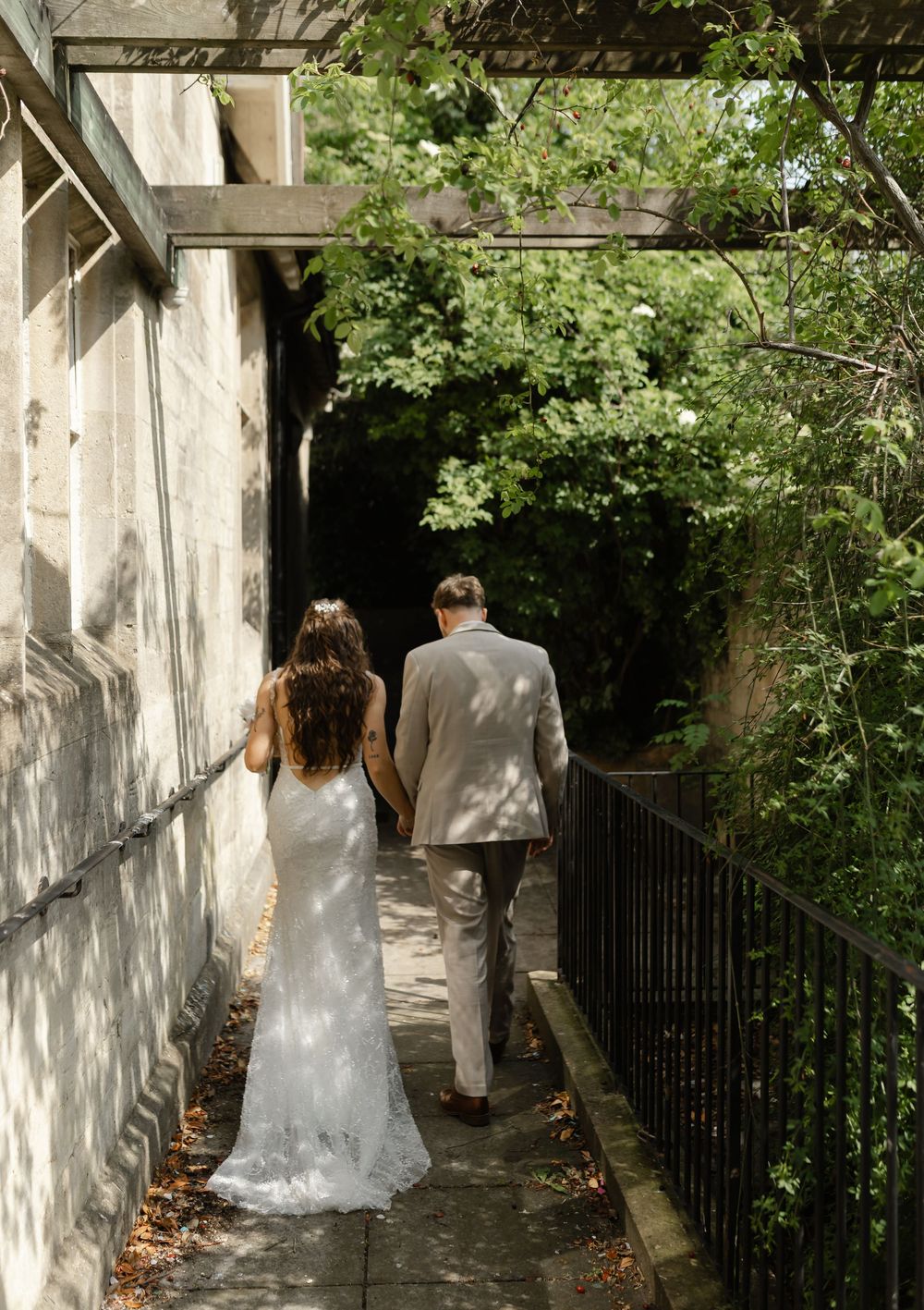 A bride and groom walk hand in hand along a sunlit garden path beside a stone wall.