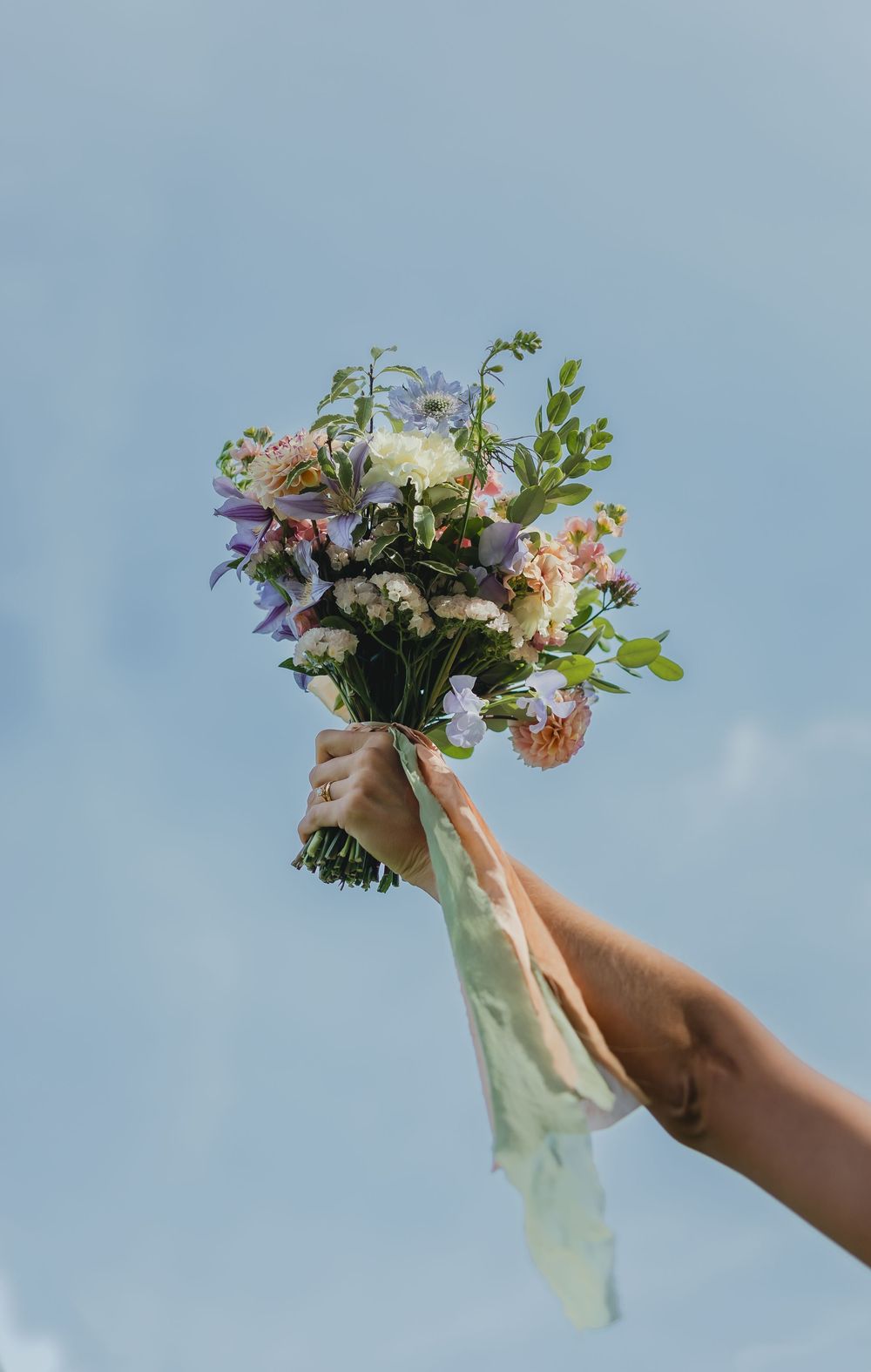Hand holding a colorful wildflower bouquet with ribbon against a blue sky.
