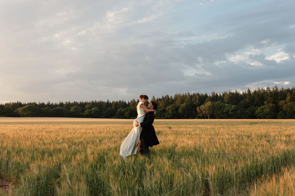Couple embracing in golden wheat field at sunset with dramatic cloudy sky and forest backdrop.