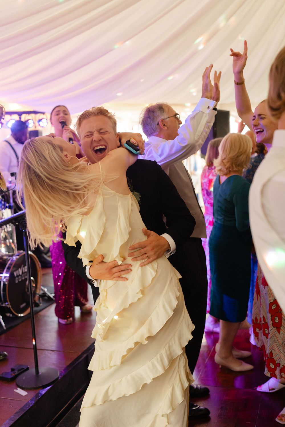 Bride in ruffled gown dances joyfully with guest at lively wedding reception under white marquee tent.