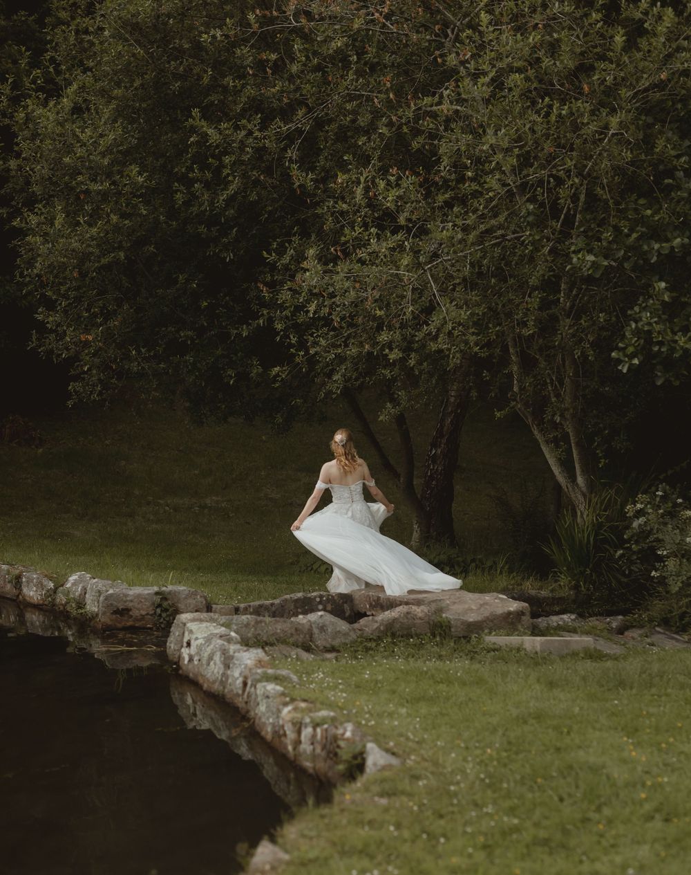 A person in a white dress sits contemplatively on rustic stone steps surrounded by lush greenery in a garden setting.