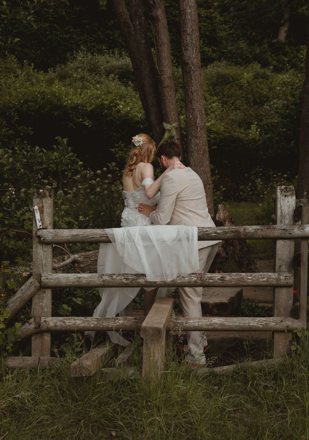 A couple in wedding attire sits intimately on a rustic wooden fence surrounded by lush greenery at sunset.