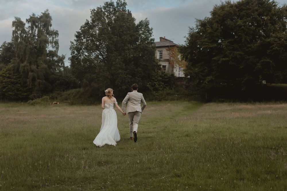 A couple in wedding attire walks through a serene meadow surrounded by trees and greenery in the afternoon light.