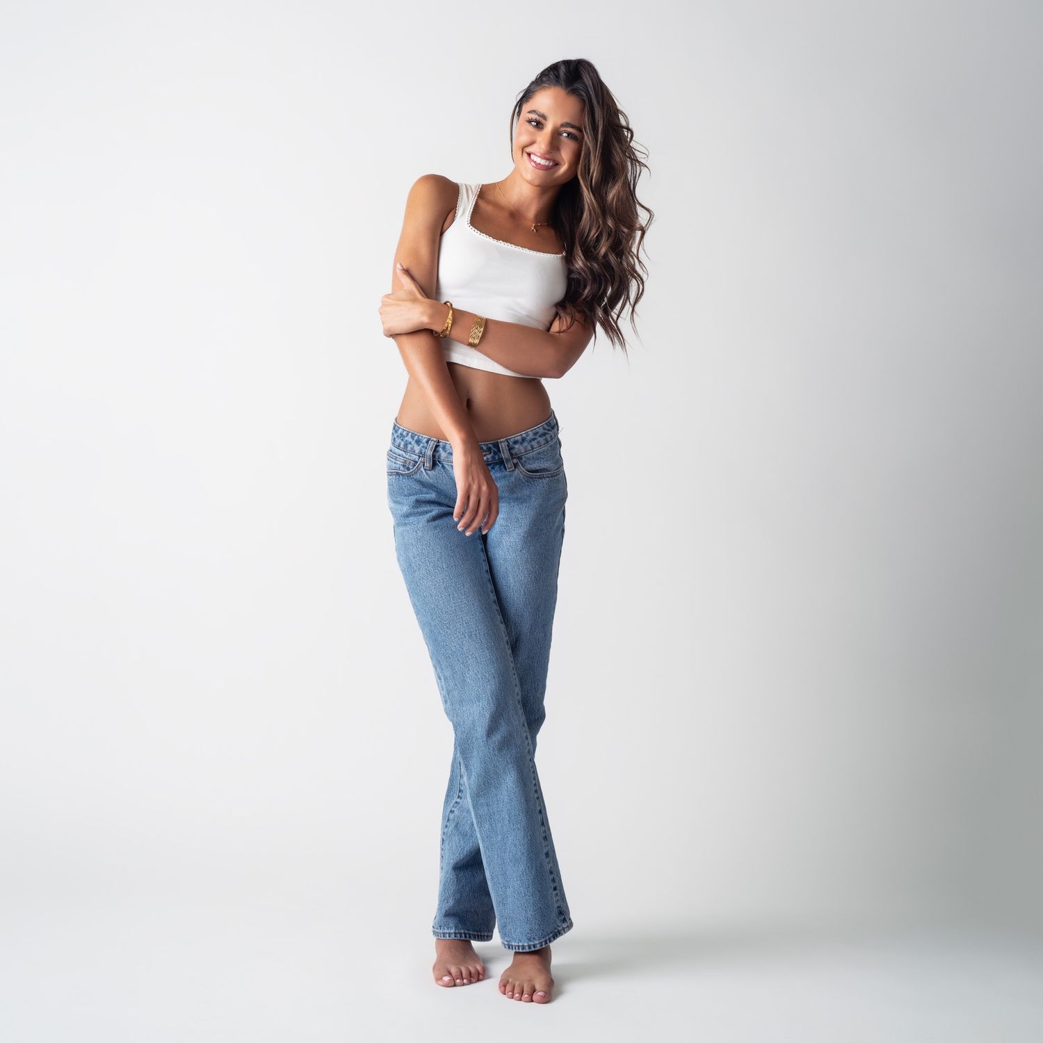 Woman in casual white crop top and light blue jeans poses against white studio backdrop in barefoot style.