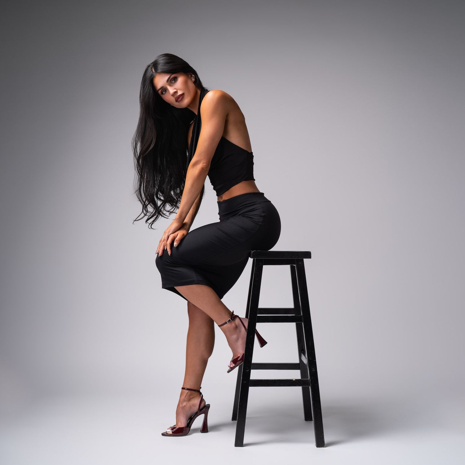 Woman in black crop top and skirt poses elegantly on wooden stool against gray studio backdrop.