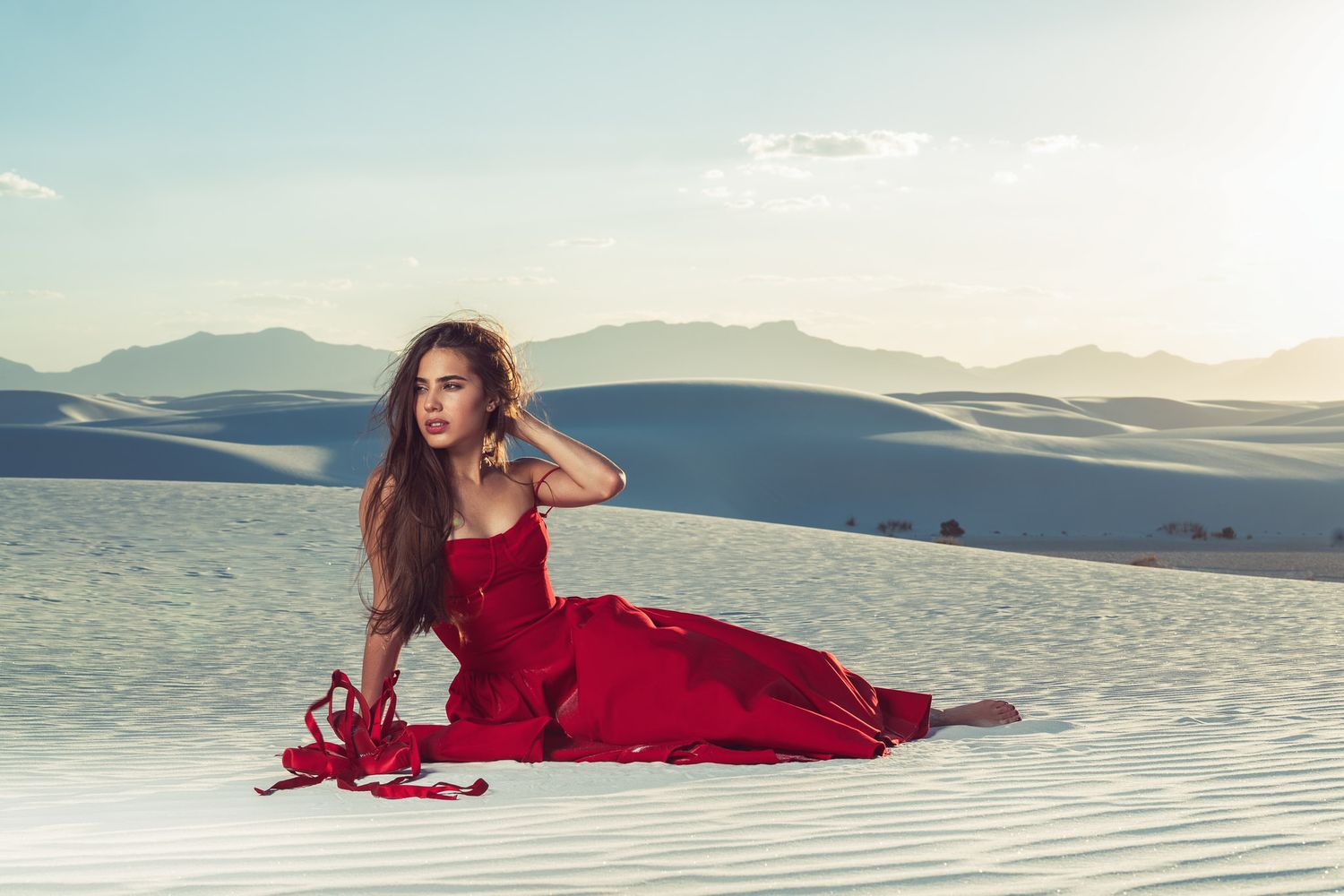 Woman in red dress posing gracefully on white sand dunes with mountains in the background.