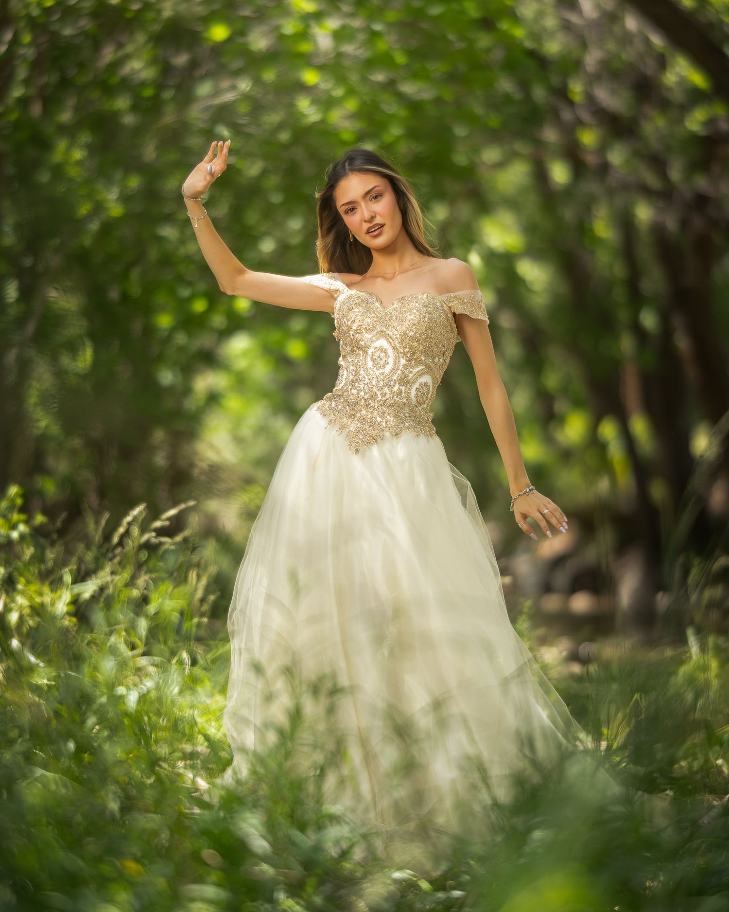 A person in an elegant off-shoulder white and gold ballgown poses gracefully in a dreamy forest setting.