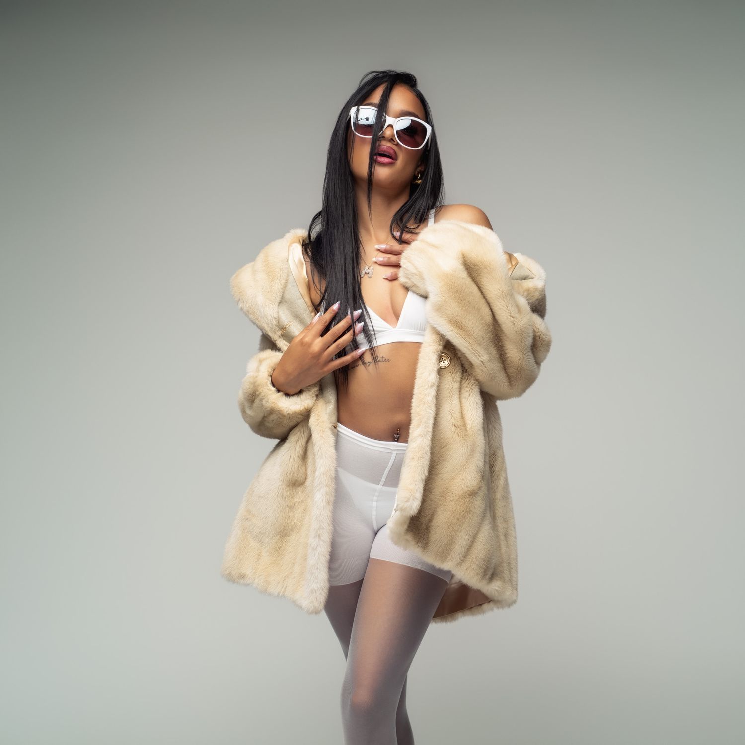 Model in cream faux fur coat and white shorts poses with sunglasses against neutral backdrop.