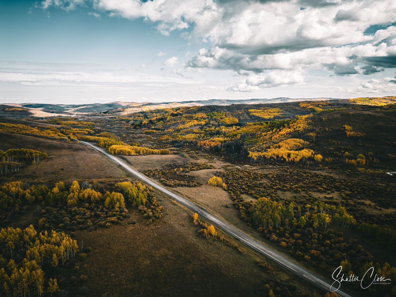 Aerial view of a winding road cutting through autumn colored hills and valleys under a cloudy sky.