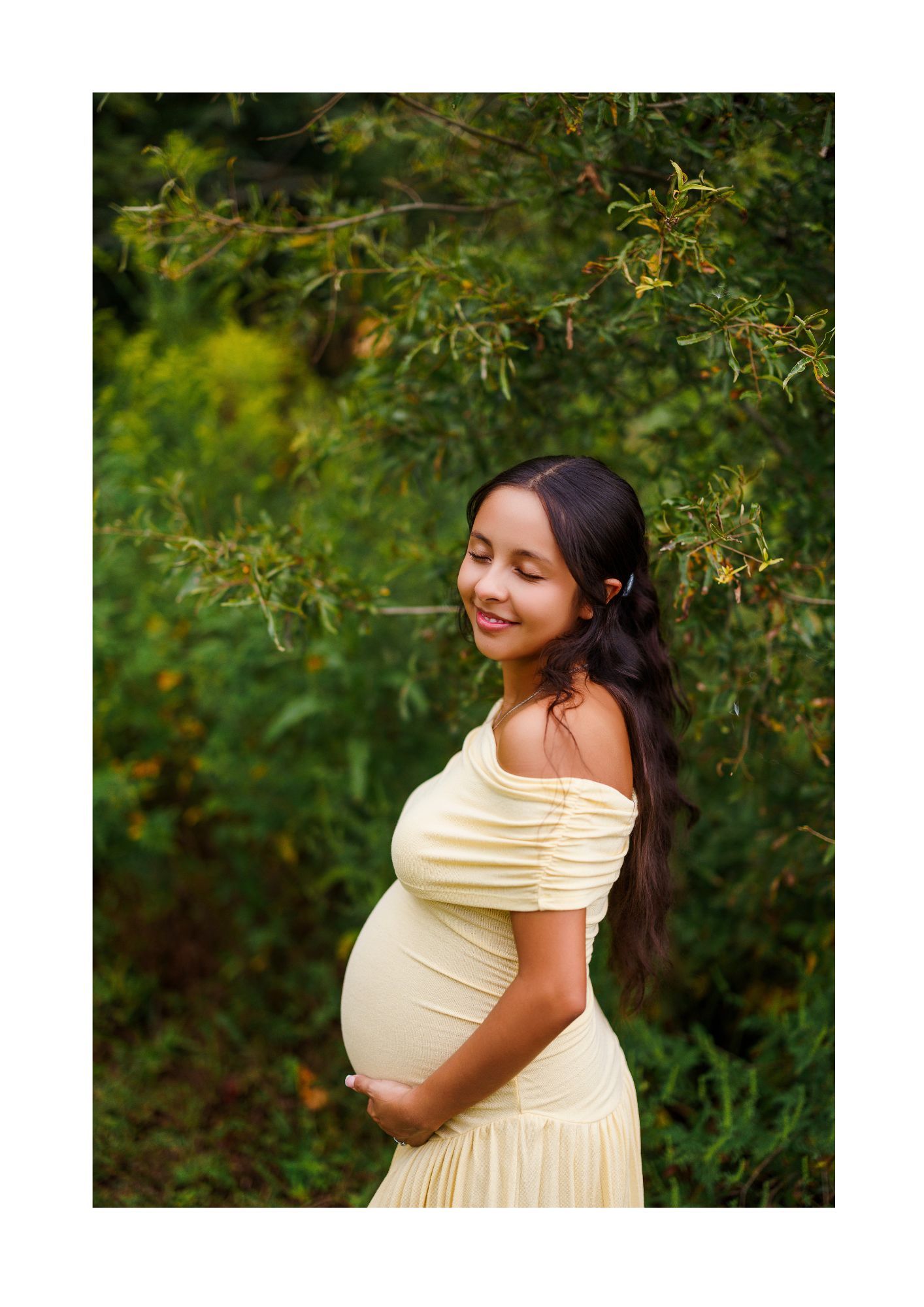 Pregnant woman in yellow off-shoulder dress posing outdoors surrounded by lush green foliage.