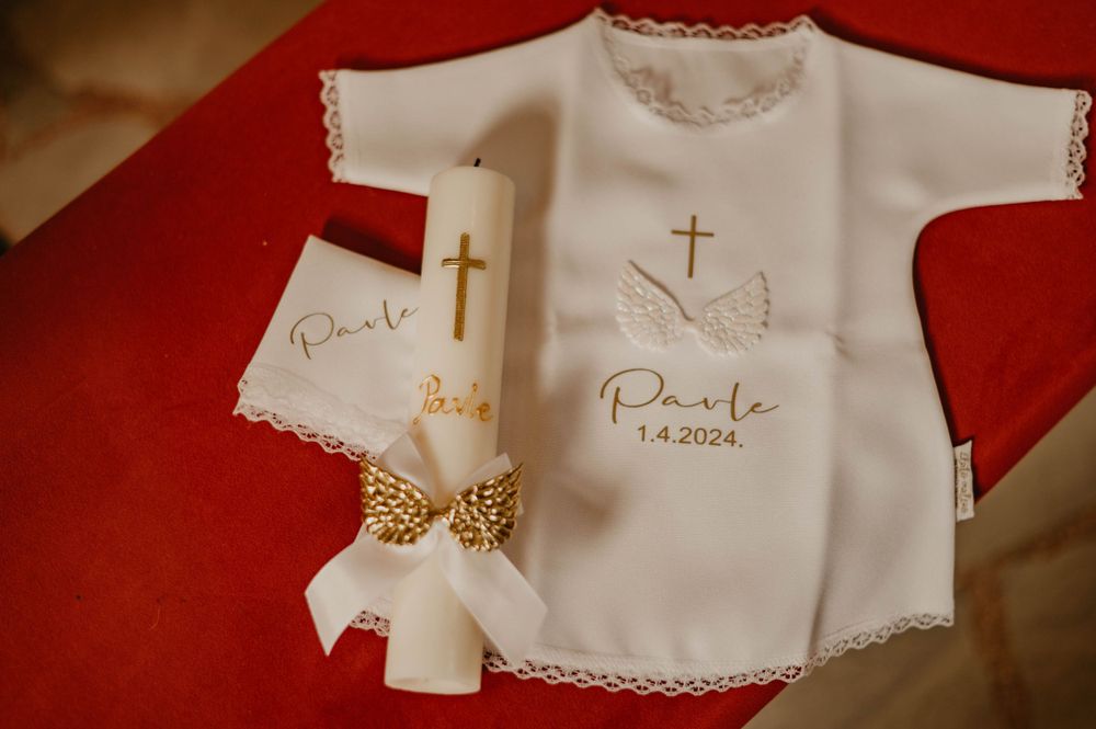 White baptismal gown with gold cross embroidery and bow detail on red background.