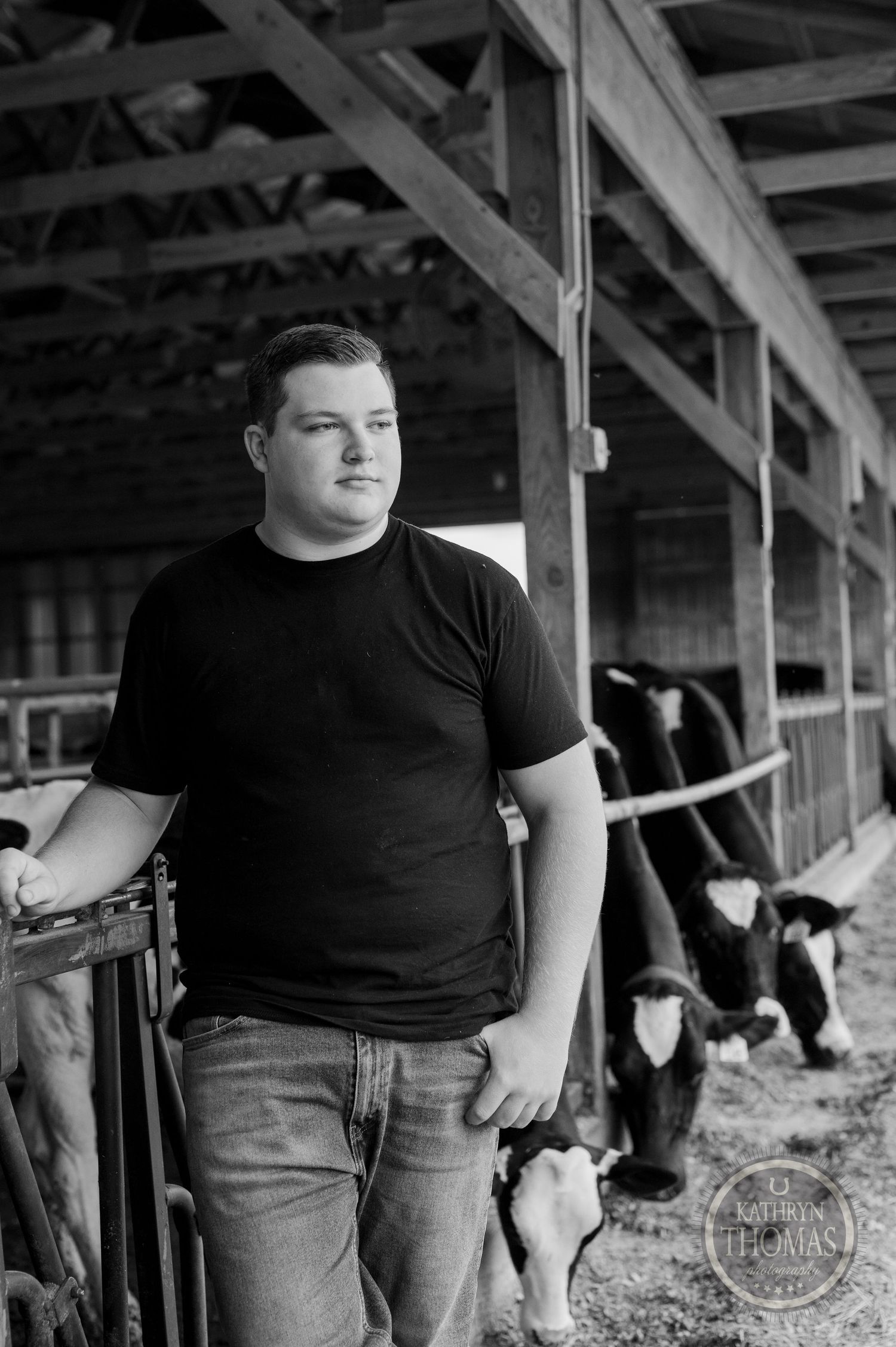 Black and white photo of someone standing in a dairy barn with Holstein cows visible in background.