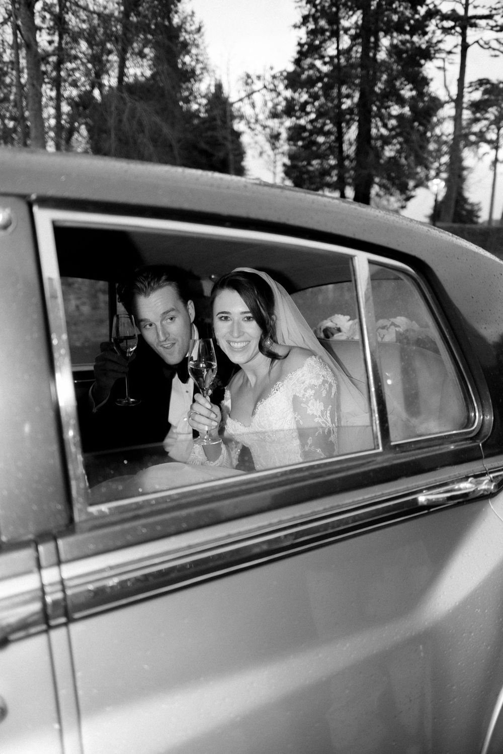 Una fotografía en blanco y negro de una pareja de recién casados ​​sonriendo desde el interior de la ventanilla de un coche clásico.