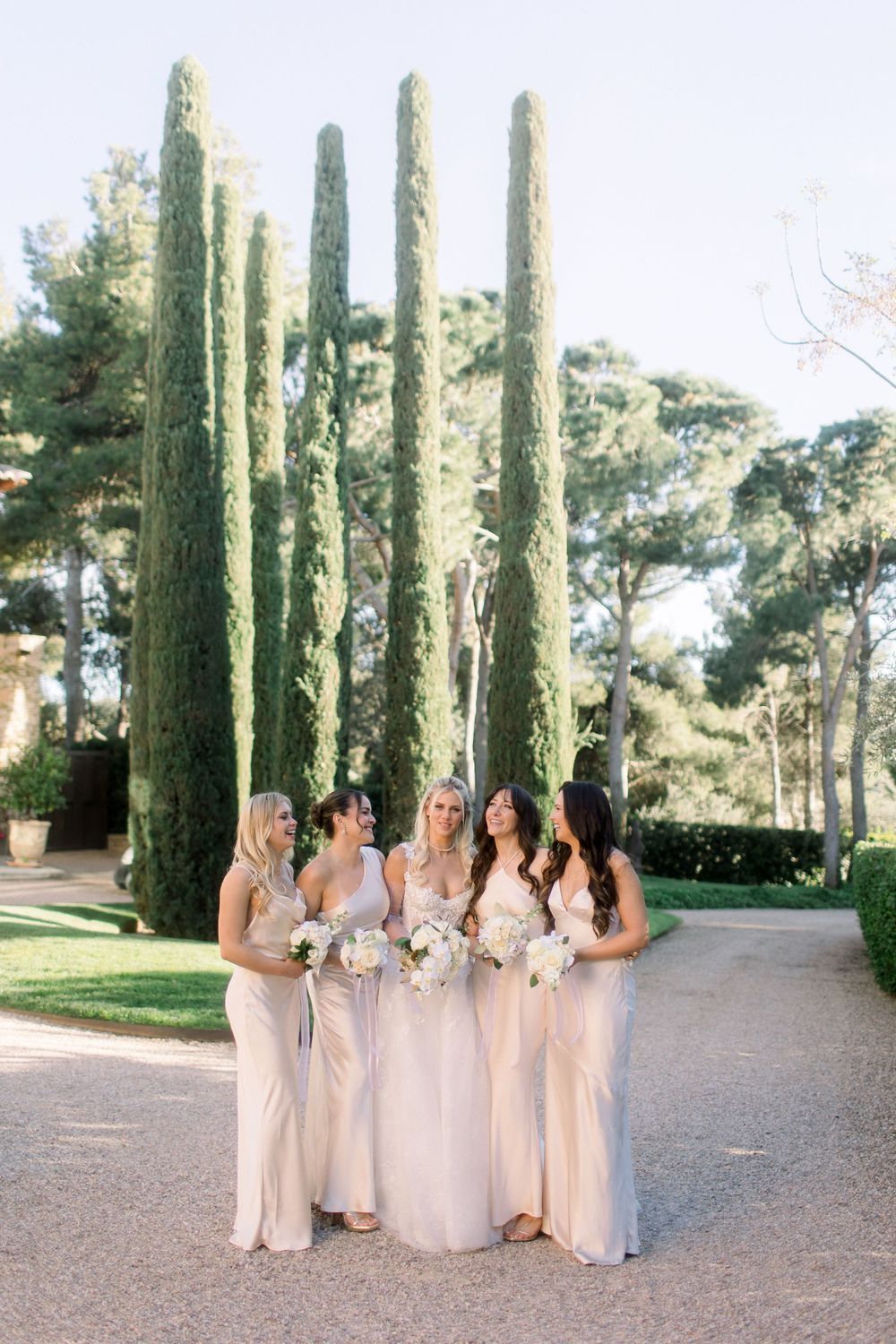 Los novios, con vestidos largos de color champán, posan juntos a lo largo de un sendero bordeado de altos cipreses en un jardín.