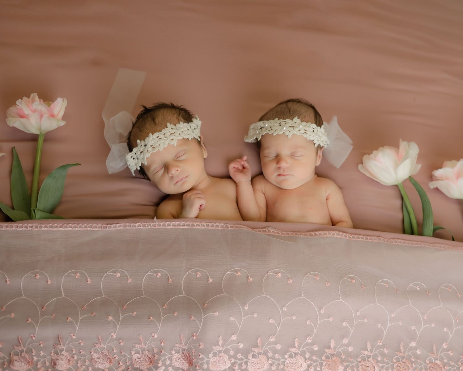Two sleeping infants lying on pink fabric surrounded by delicate tulip flowers for a newborn photoshoot.
