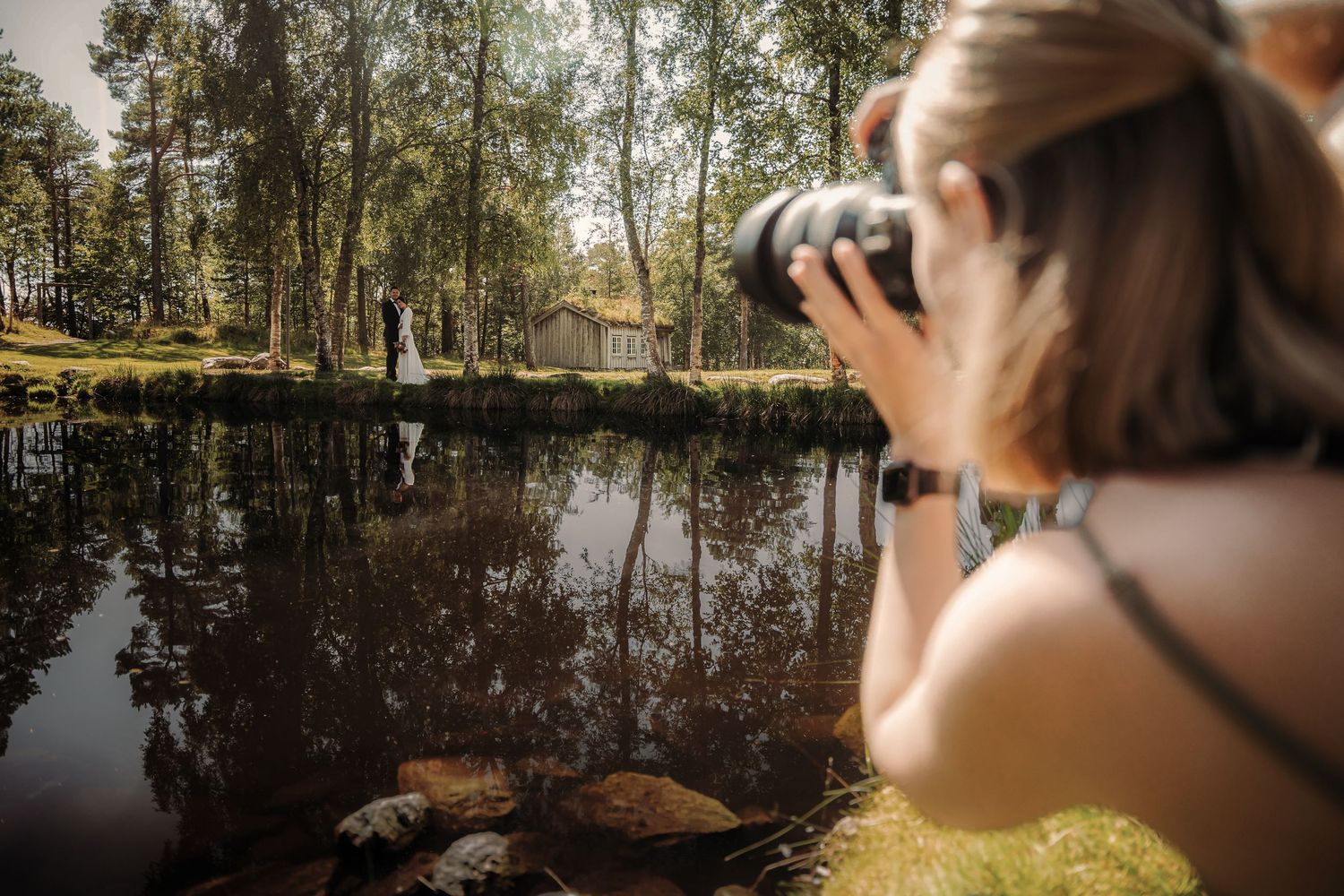 Photographer taking nature shots at tranquil pond with trees reflecting in still water.