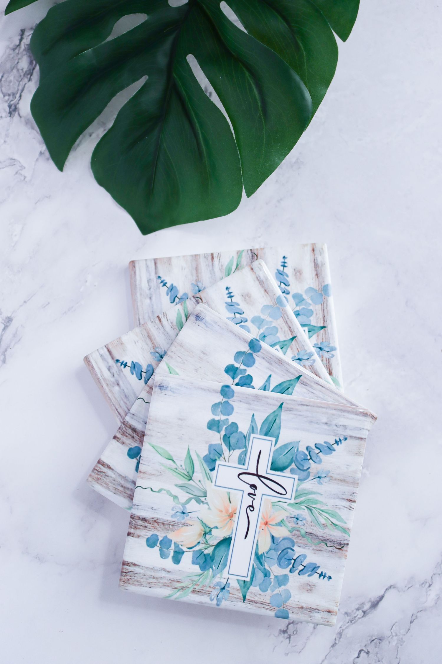 Decorative cards with blue floral designs arranged under monstera leaves on marble background.