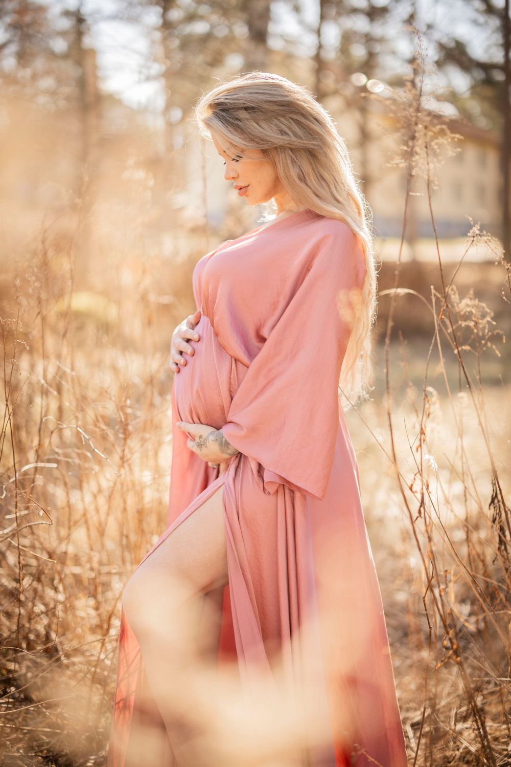 Maternity portrait in pink flowing dress against sunlit autumn field with soft bokeh effect in background.