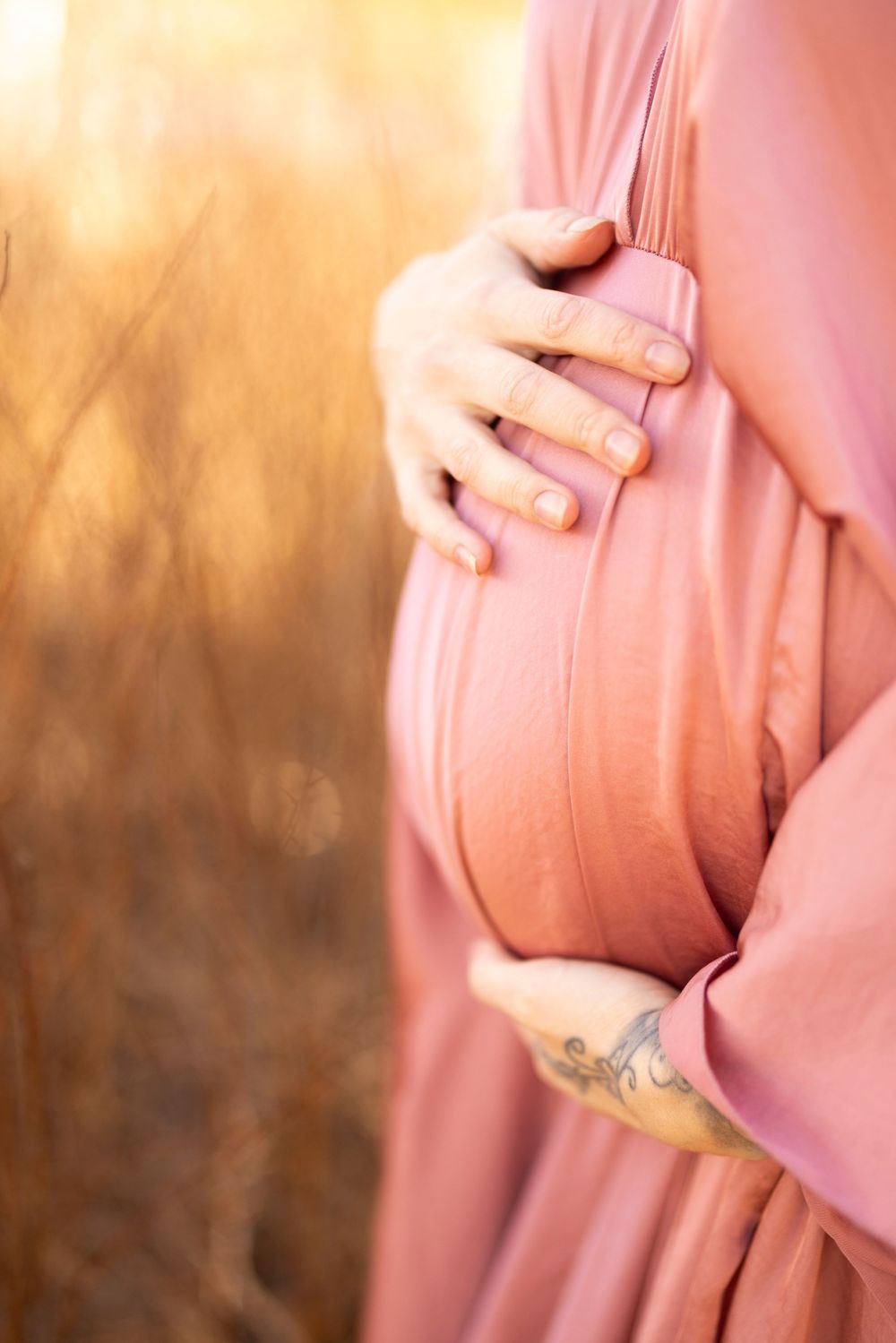 Close-up of pregnant woman in flowing pink dress holding her baby bump against golden sunset lighting.