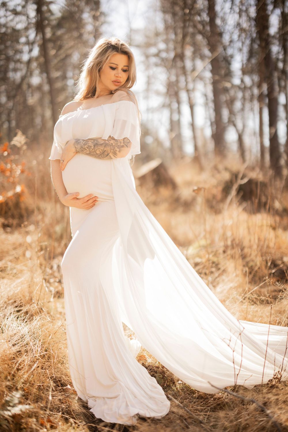Ethereal maternity portrait of a woman in a flowing white gown standing in golden autumn grass at sunset.