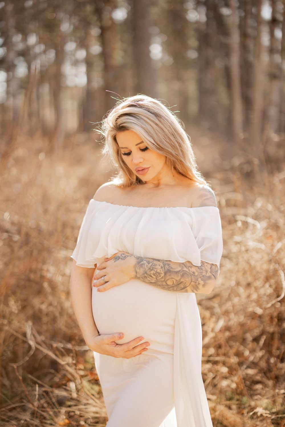 An expectant mother in a flowing white dress caresses her belly while standing in a golden sunlit forest.