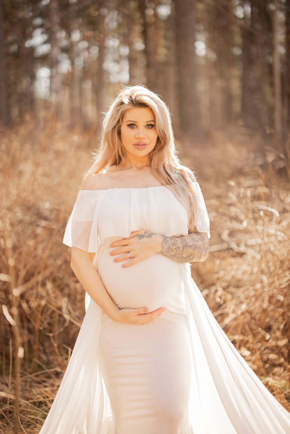 Woman in flowing white off-shoulder dress poses in autumn forest during golden hour maternity photoshoot.