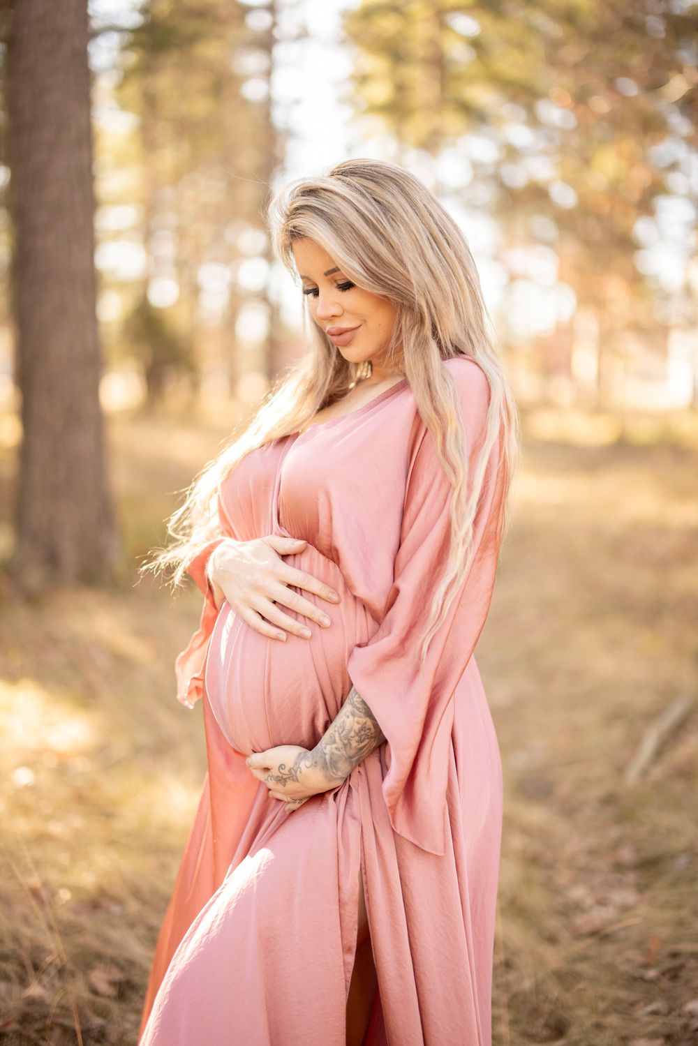 Expectant person in flowing pink dress cradles baby bump during outdoor maternity photoshoot in golden sunlit forest.