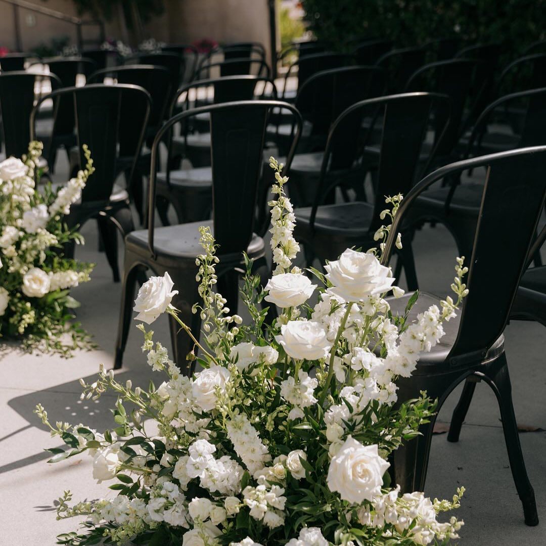 Elegant white floral arrangements with roses and greenery alongside black chairs at wedding ceremony setup.