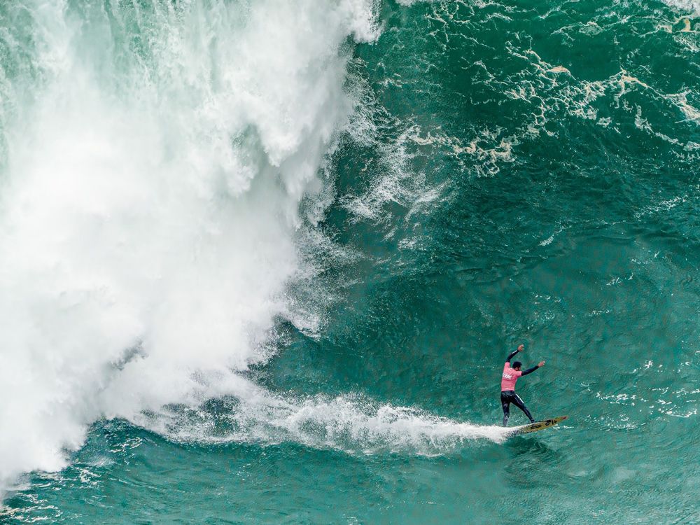 Aerial view of surfers riding massive turquoise waves in powerful ocean surf conditions.