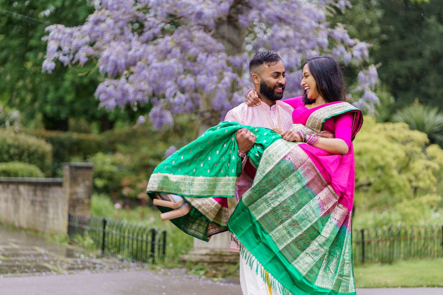 A couple in traditional South Asian attire dance together in front of blooming purple wisteria in a garden setting.