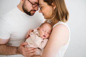 Intimate family portrait with parents embracing their newborn baby against white backdrop.
