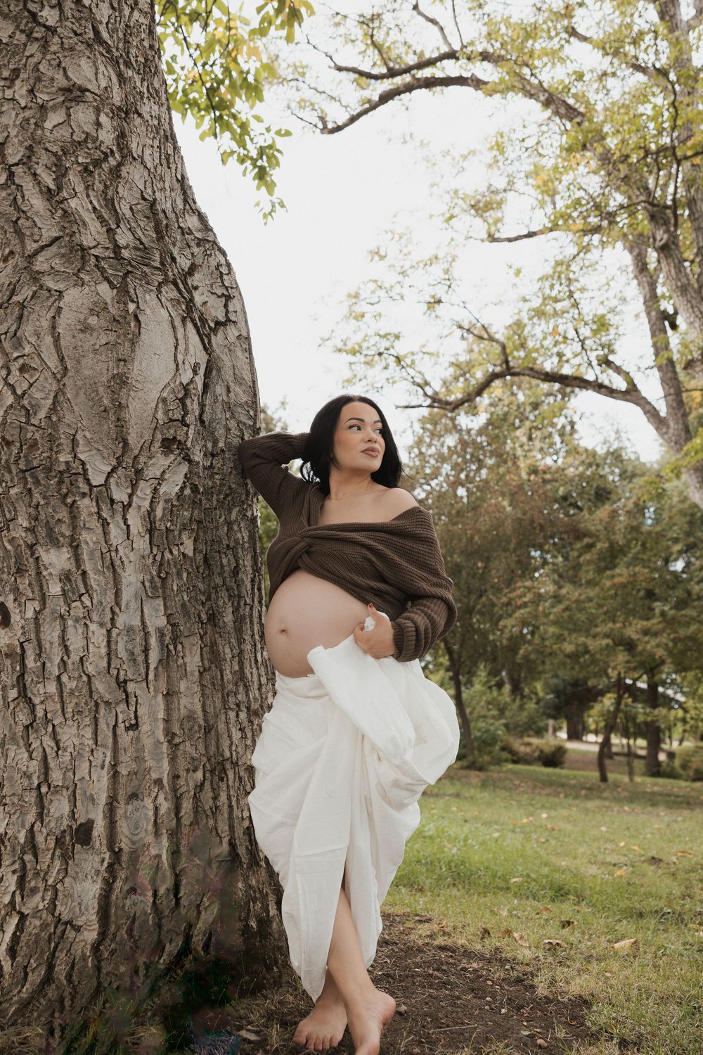A figure in a flowing white dress poses gracefully against a tree trunk in a sunlit outdoor setting.