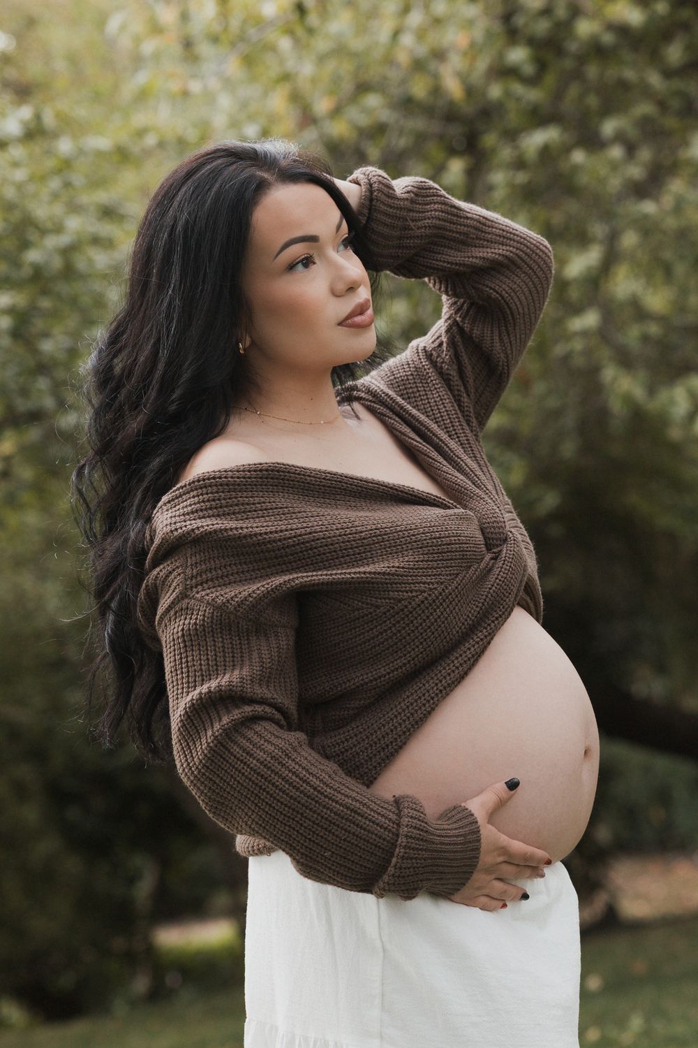 A maternity photoshoot in a garden setting shows a person wearing a brown sweater and white dress cradling their belly.
