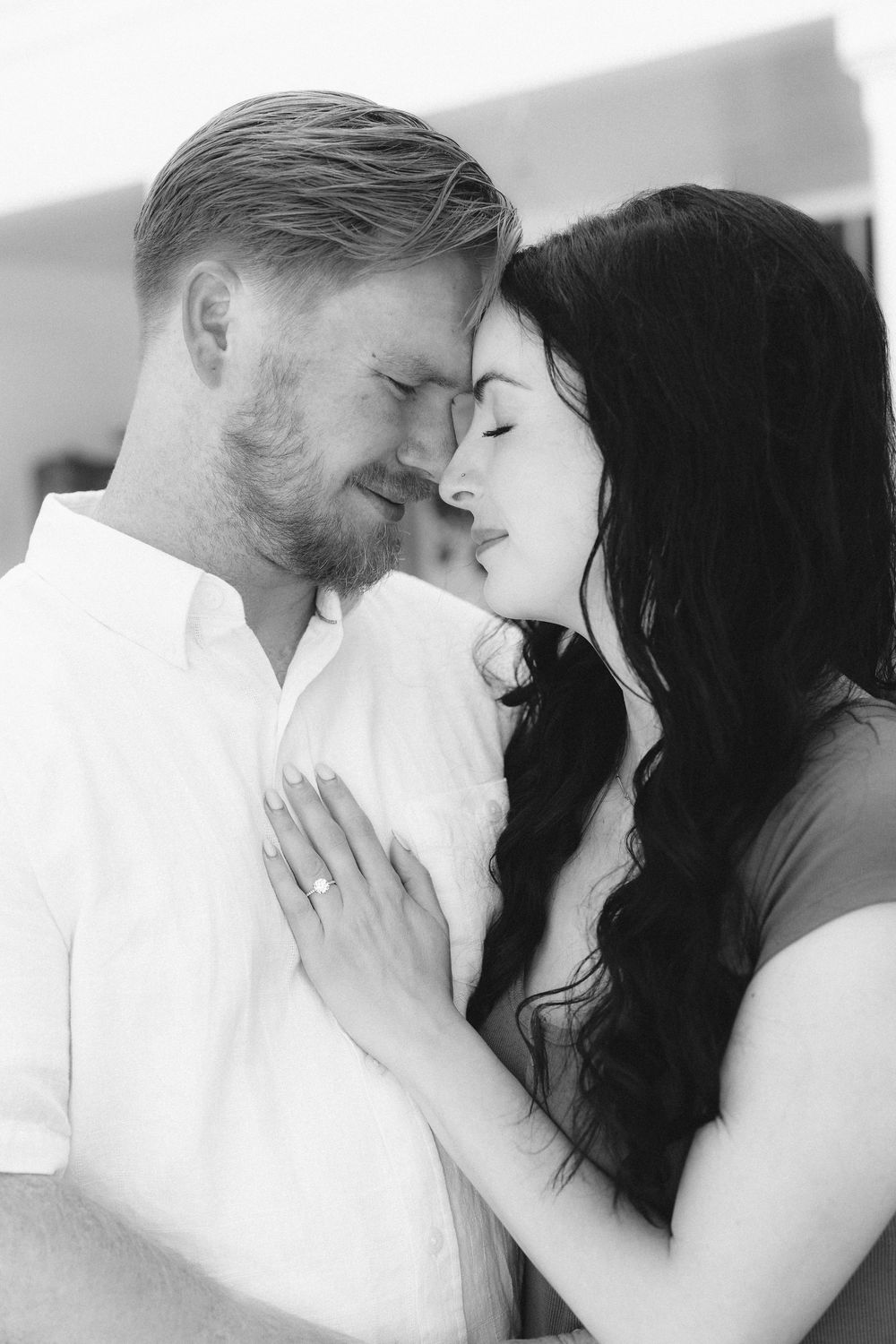 Black and white photo of a romantic couple with foreheads touching, woman wearing engagement ring resting hand on mans chest.