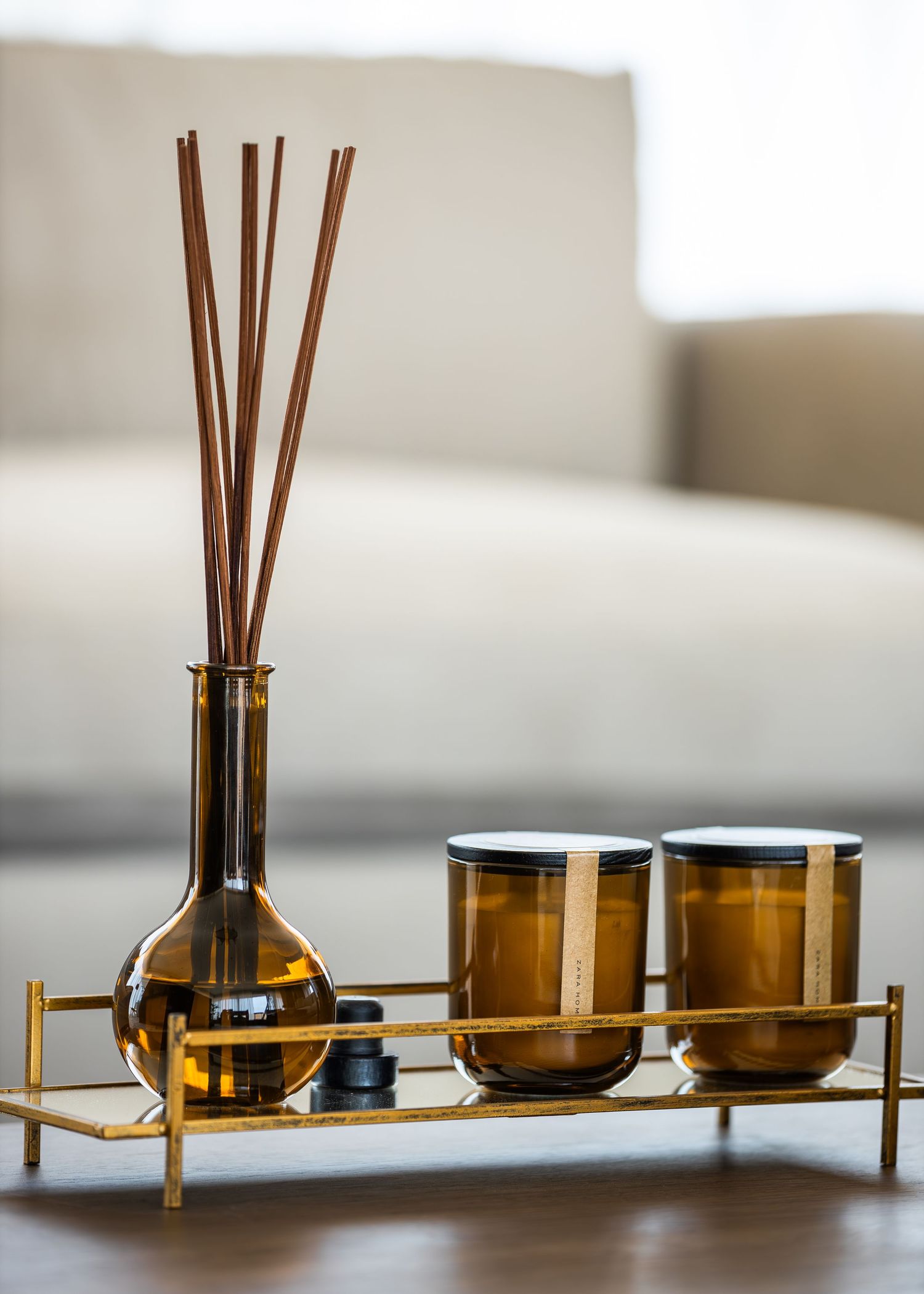 Reed diffuser with amber glass bottle and two matching candles arranged on a sleek gold metal tray.