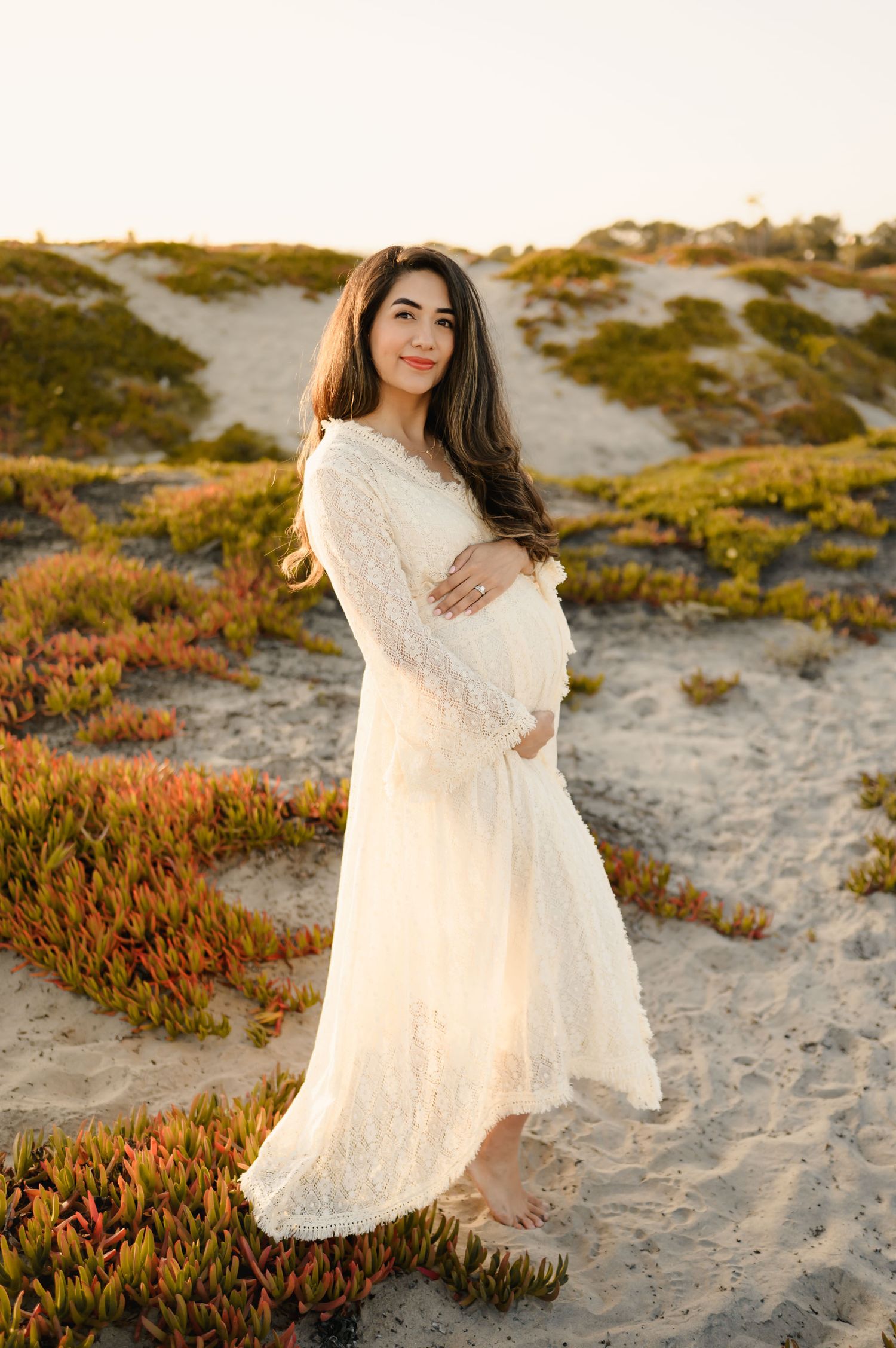 woman in see through lace dress holds pregnant belly during her sunset beach photoshoot by Ventura and Santa Barbara Photographer