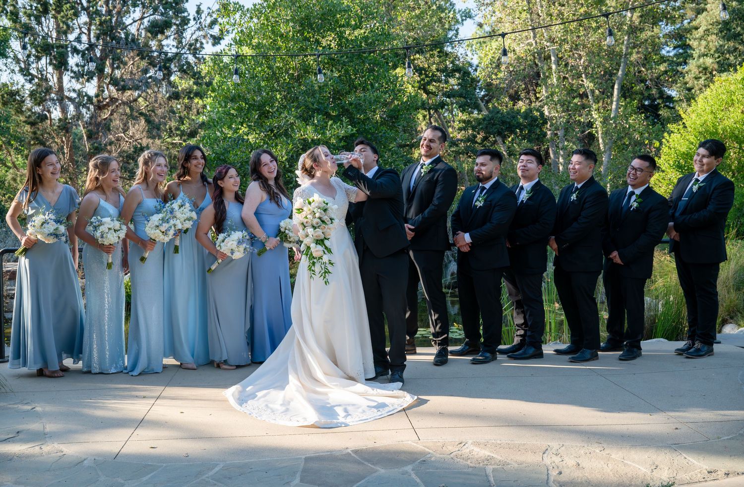 A wedding party in light blue bridesmaid dresses and black suits poses outdoors on a sunny day.