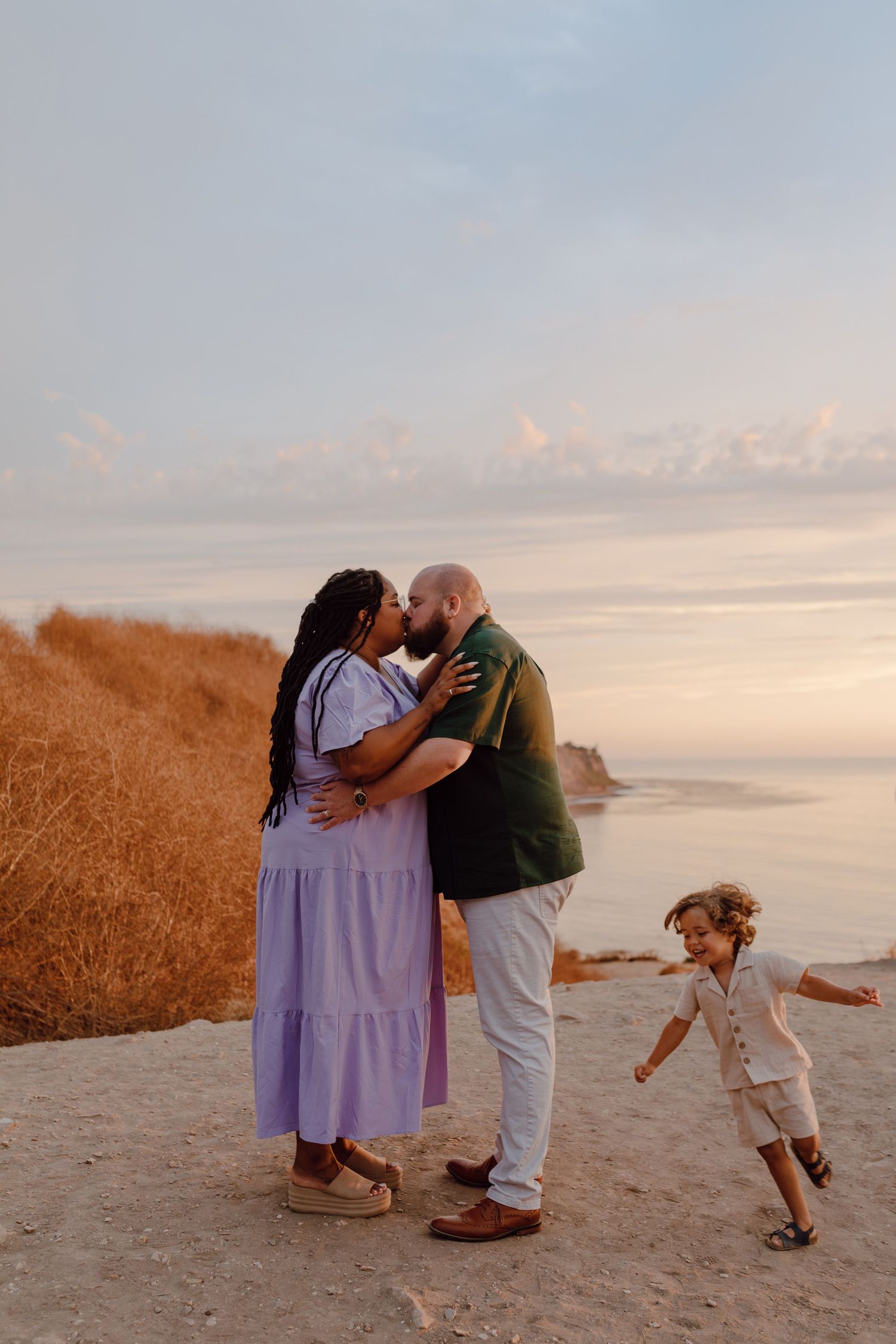 A sweet moment shared on a coastal cliff at sunset while a child plays nearby.