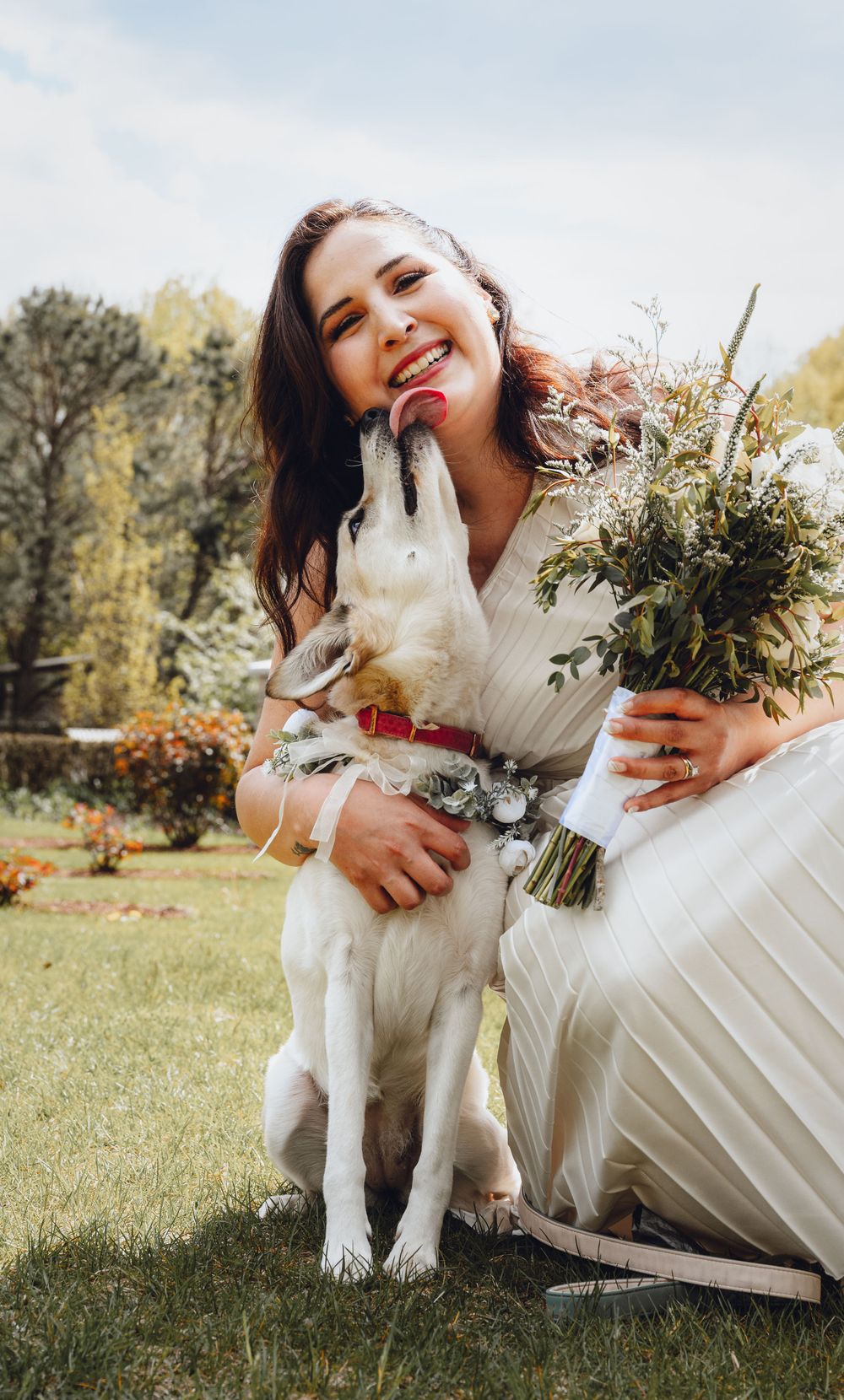 White dog wearing floral collar poses with wedding bouquet outdoors.