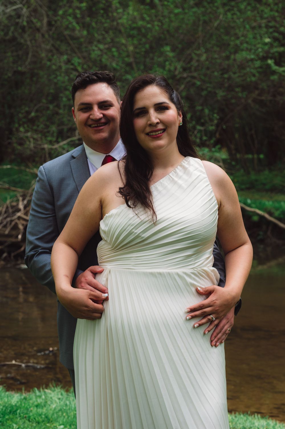 Romantic portrait of newlyweds in formal attire by a stream.
