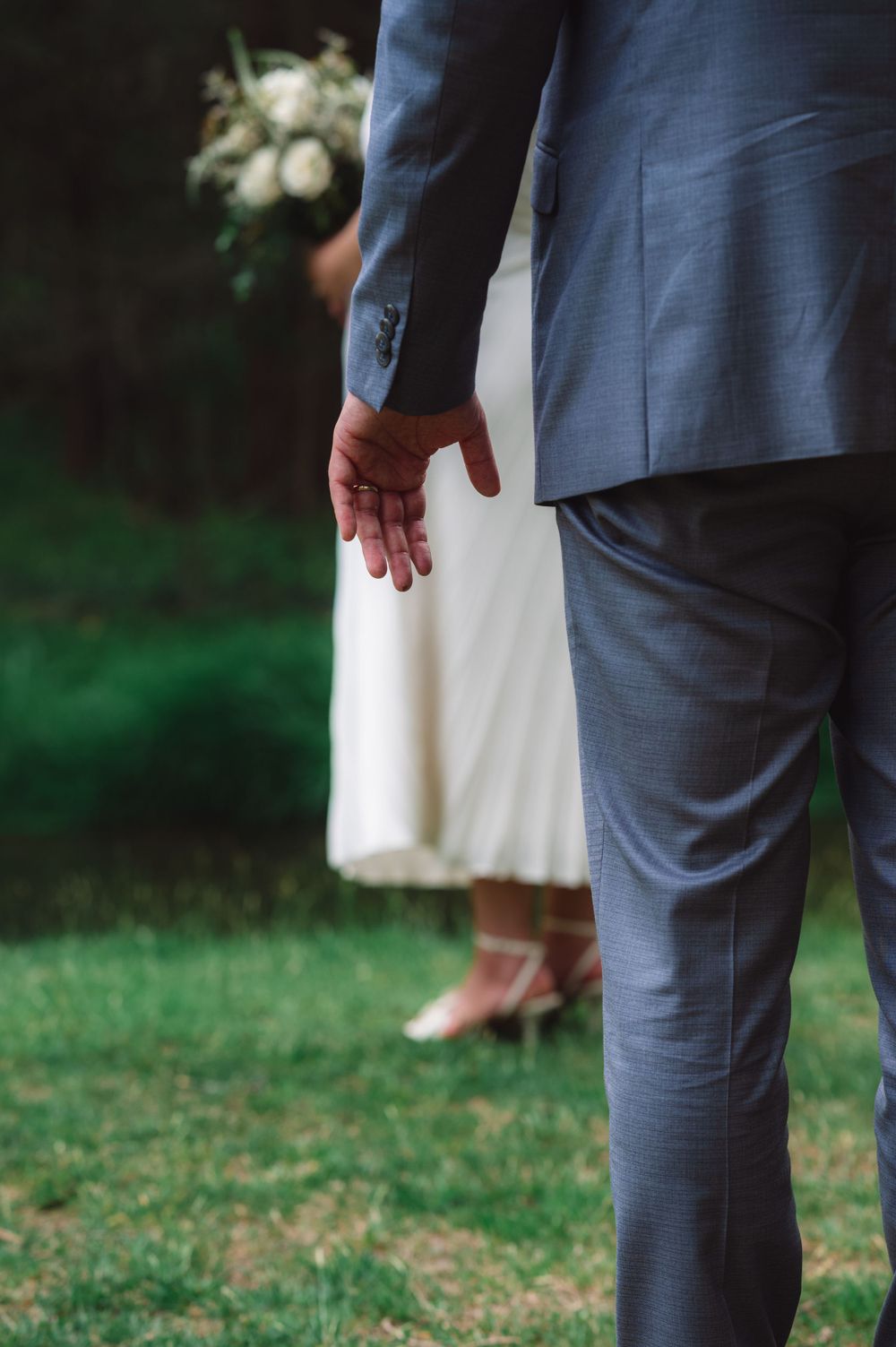 Detail shot of formal wedding attire with gray suit and white dress against green grass.