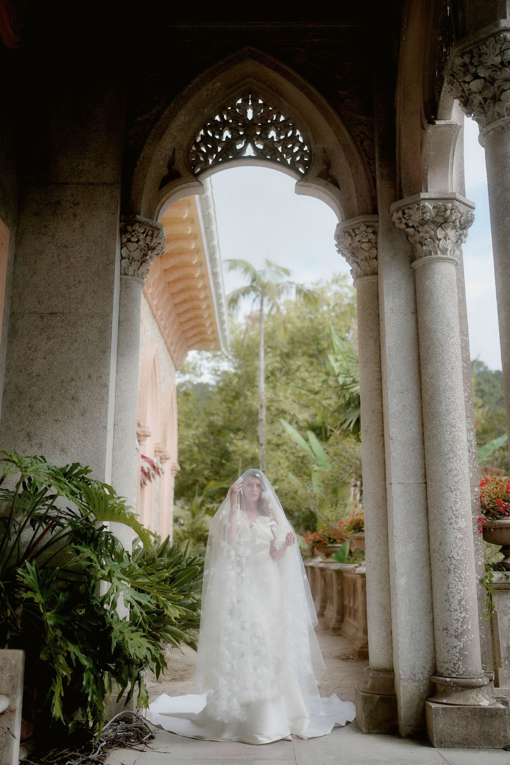 Bride in flowing white wedding dress and veil poses elegantly under ornate stone archways surrounded by tropical foliage.