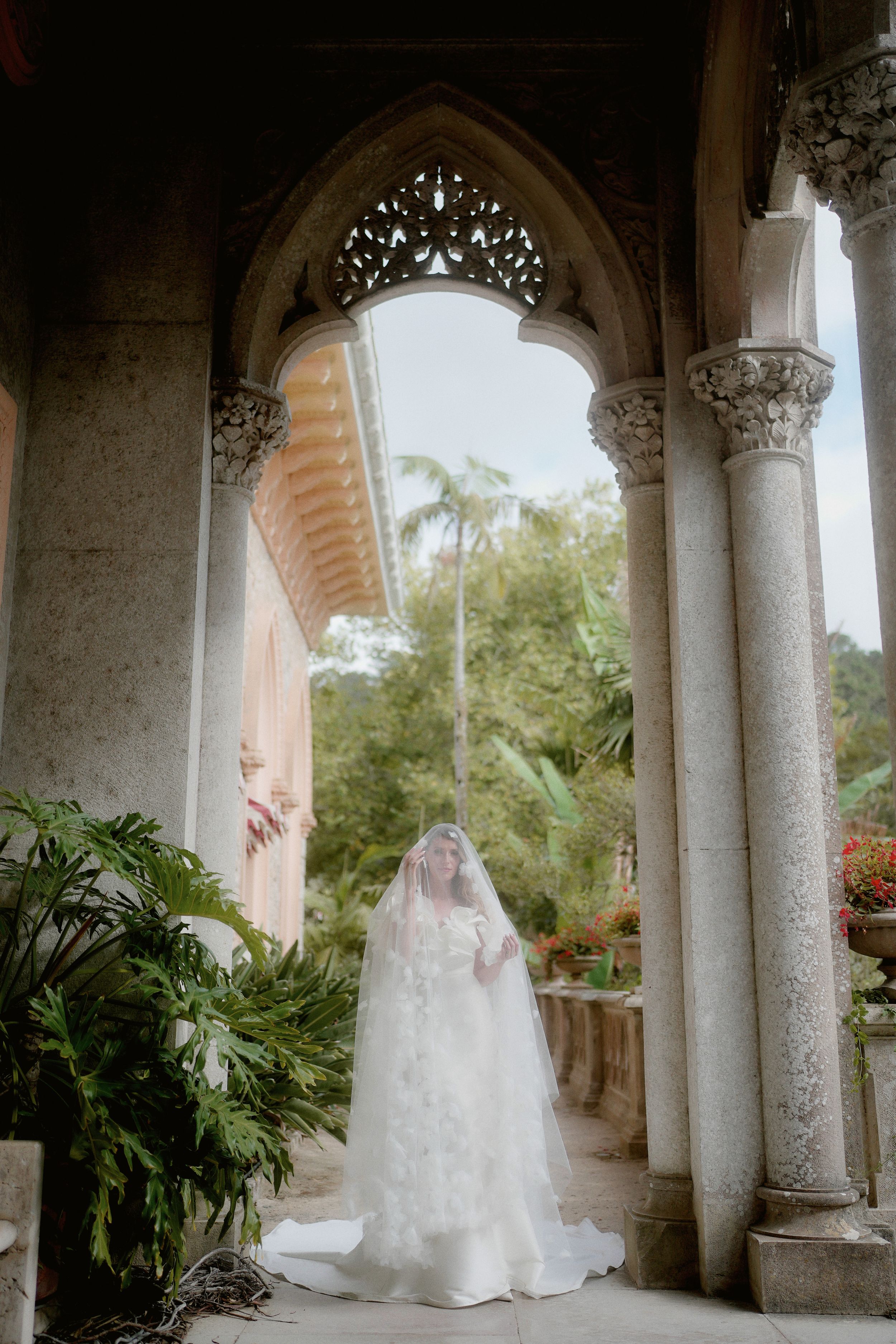 Bride in flowing white wedding dress and veil poses elegantly under ornate stone archways surrounded by tropical foliage.