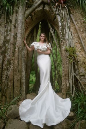 Elegant white wedding gown with mermaid silhouette and ruffled off-shoulder detail photographed in stone archway with tropical foliage.